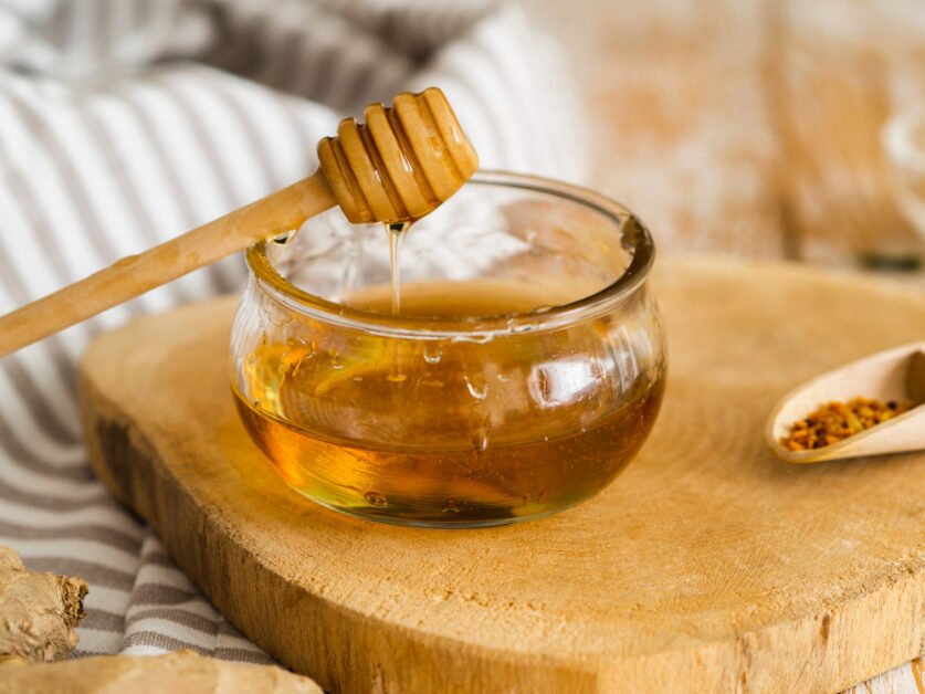 Glass bowl filled with honey on a wooden piece, with a wooden honey spoon resting on the edge and honey dripping into the bowl