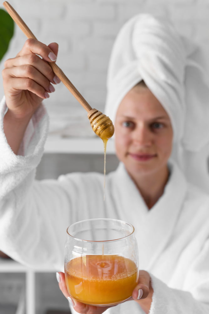 Woman in a bathrobe with rolled hair holding a glass bowl half-filled with honey and a wooden spoon with honey dripping into the bowl.
