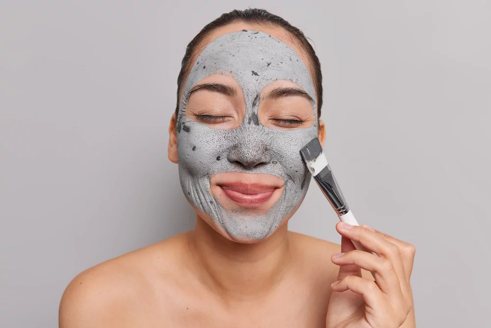 Woman with combed hair applying a mask to her face with a cosmetic brush, standing shirtless indoors.