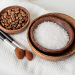 Wooden bowls with coffee beans and sugar next to cosmetic brushes on a white towel, illustrating ingredients for the best face exfoliator scrub.