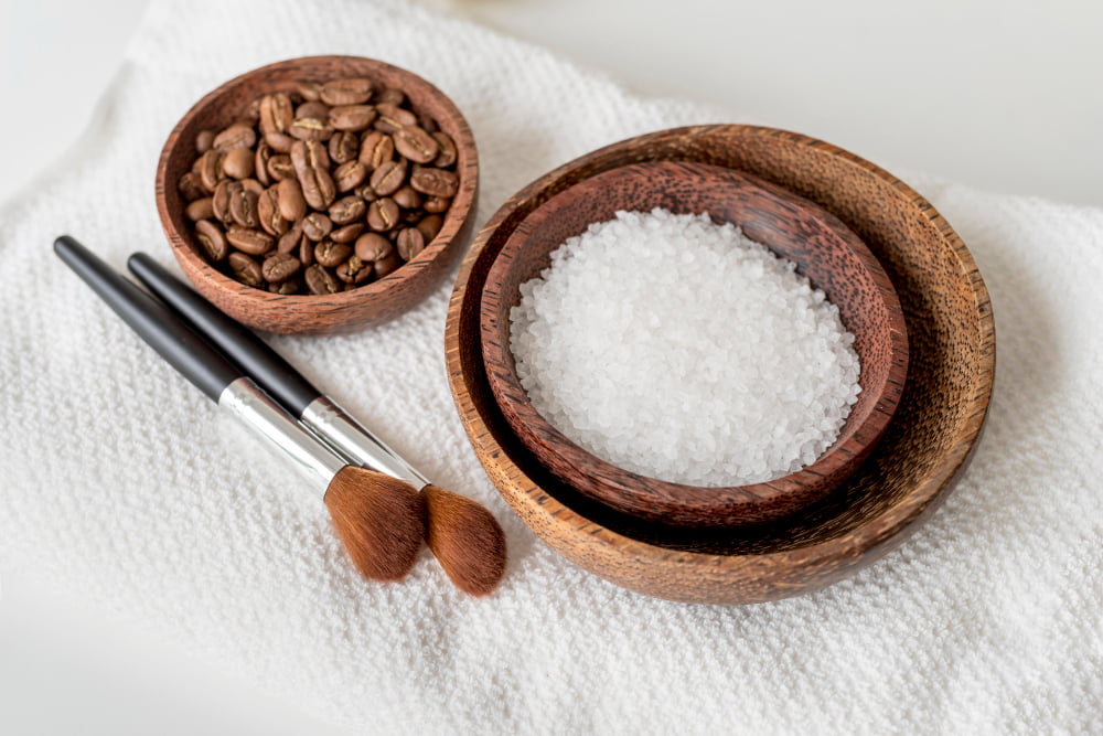 Wooden bowls with coffee beans and sugar next to cosmetic brushes on a white towel, illustrating ingredients for the best face exfoliator scrub.